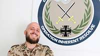 Ein Soldat steht lachend vor dem Kontingentwappen mit der Aufschrift „Operation Inherent Resolve“