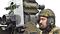 Ein Soldat mit Sprechhaube blickt durch den Sucher. Links ist die Zieloptik der Panzerabwehrwaffe.
