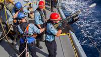 Ein Soldat auf einem Schiff zielt mit einem Gewehr aufs Meer, drei Soldaten stehen hinter ihm.