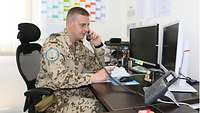 Ein Soldat sitzt an einem Schreibtisch mit zwei Monitoren und telefoniert
