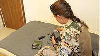 Eine graue Decke ist über ein Bett gespannt, darauf sitzt eine Soldatin, die eine zerlegte Pistole reinigt