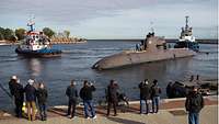 Ein U-Boot in einem Hafen, kurz vor einer Pier, zwei Schlepper dahinter.