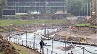 Eine Kanuslalom-Wildwasserstrecke verläuft mitten durch das Bild. Im Hintergrund ist ein Gebäude zu sehen, das umgebaut wird.