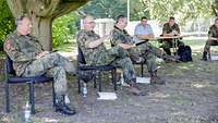 Drei Soldaten sitzen auf Stühlen unter einem Baum mit Abstand nebeneinander, ein Soldat spricht.