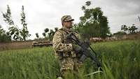 Ein Soldat mit einer Waffe steht in einem grünen Kornfeld, im Hintergrund ein Gefechtsfahrzeug.