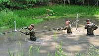 Drei Soldaten stehen bis zur Brust im Wasser und ziehen sich watend an einem Seilsteg entlang.