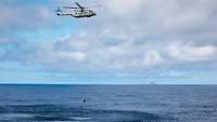 Ein Hubschrauber zieht am langen Draht einen zylindrischen Körper aus dem Wasser. Im Hintergrund eine dänische Fregatte.