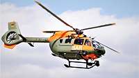 Ein Hubschrauber fliegend, oliv-orangefarben lackiert. Auf der Tür in Weiß SAR für Search and Rescue