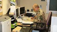 Ein Soldat sitzt in einem Büro, vor ihm verschiedene Papiere und zwei Computermonitore