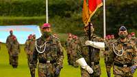Drei Soldaten marschieren nebeneinander, der mittlere trägt die schwarz-rot-goldene Truppenfahne.