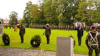 Vier Soldaten der Bundeswehr stehen, militärisch grüßend, vor vier Blumenkränzen und einem Gedenkstein.
