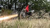 Zwei Soldaten hocken neben einem feuernden Raketen-Panzerabwehrsystem im Gras. 