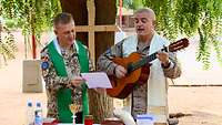 Zwei Militärpfarrer singen, einer spielt Gitarre. Im Hintergrund ein Baum mit Holzkreuz, vorne ein Tisch als Altar. 