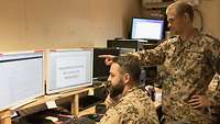 Zwei Soldaten in einem Raum mit mehreren Computern, der eine sitzt am PC, der andere steht und weist auf einen Bildschirm