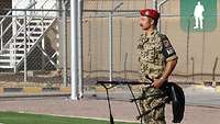 Ein Soldat hält einen Dudelsack in der linken Hand und steht auf dem Sportplatz, im Hintergrund eine Mauer mit Turm