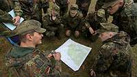 Mehrere Soldaten stehen und hocken im Gelände um eine Karte herum. Einer von ihnen hält einen Kompass in der Hand und spricht.