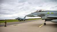 Ein britischer und ein deutscher Eurofighter rollen gemeinsam auf dem Flugplatz in Ämari. Ein Einwinker weist den Jets den Weg.