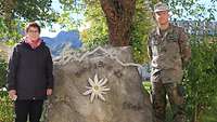 Eine Frau in Zivil und ein Mann in Uniform stehen links und rechts neben einem großen Stein.