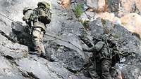 Zwei Soldaten mit Gepäck und Waffen klettern an einer Felswand hinauf.