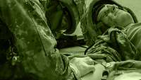 A medical orderly applies an intravenous catheter to the arm of an injured fellow soldier lying on the ground.