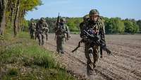 Five soldiers move in column along the edge of a field. In lead position is a medic.