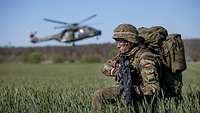 A soldier kneels in a verdant cornfield, as a helicopter lands in the background.