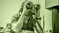 A soldier uses binoculars to observe from within a vehicle.