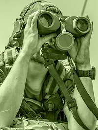 A soldier uses binoculars to observe from within a vehicle.