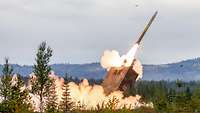A rocket launcher positioned in a forest area launches a missile, leaving a fire trail and smoke.