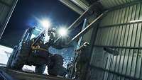 A wheel loader drives up a ramp into a loading bay. A soldier stands to one side and guides the vehicle.