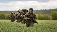 Six armed paratroopers move in single file through a green field of grain.