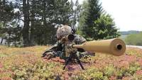 A soldier with a sniper rifle lies in vegetation-covered terrain.