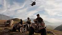 Soldiers, two quads, a motorbike and two off-road vehicles against the backdrop of a mountain range and a helicopter in the sky.