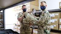 Zwei Soldaten mit Mund-Nasen-Schutz stehen in einem Lagerraum und halten gemeinsam eine Schutzweste in den Händen