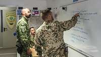 Ein deutscher, ein kroatischer und ein ungarischer Soldat schauen auf ein Whiteboard, während der Deutsche auf etwas zeigt