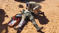 Ein Soldat versorgt einen Verwundeten in der Wüste, im Hintergrund ein VN-Fahrzeug 
