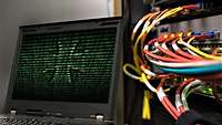 Symbolfoto: Laptopbildschirm mit Schrift und einem Eisernen Kreuz in Grün, daneben ein Serverschrank mit farbigen Kabeln.