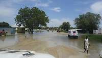 Mehrere Fahrzeuge fahren auf einer überschwemmten Straße, im Hintergrund Container und Bäume, vorn eine Einheimische 