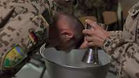 Ein Soldat hält den Kopf über eine Schüssel, eine Hand hält den Kopf und gießt aus einem Kelch Wasser darüber