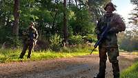 Zwei Soldaten stehen mit dem Gewehr in der Vorhalte links und rechts eines Waldweges.