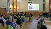 Ein Soldat steht in einem großen Saal vor den mit Abstand sitzenden Teilnehmenden und hält einen Vortrag