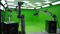 Three robot arms with cameras stand in front of a green wall in a room