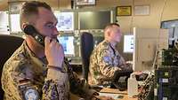 Zwei Soldaten sitze am Schreibtisch vor ihren Computern. Ein Soldat hält einen Telefonhöhrer a seinem Ohr.