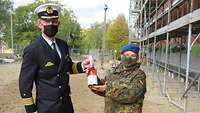 Ein Mann in Marineuniform übereicht einer Frau in Flecktarnuniform eine Flasche Sekt