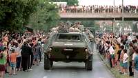 Viele Menschen stehen am Straßenrand und winken vorbeifahrenden Soldaten in einem Panzer zu