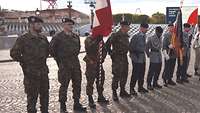 Soldaten verschiedener Nationen stehen auf einem Platz in Abständen, zwischen ihnen Truppenfahnen.