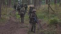 Eine Gruppe von Soldaten marschiert mit Gepäck eine Anhöhe im Wald hinauf, ein Soldat dreht sich um.