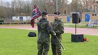 Zwei Soldaten stehen auf dem Rasen eines Sportplatzes. Ein Soldat überreicht einem anderen einen schwarz-roten Militärwimpel.