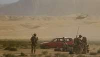 Ein Auto steht in der Wüste, Soldaten stehen und knien davor und vor dem Gebirge im Hintergrund fliegt ein Hubschrauber