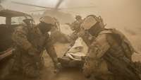 Ein Soldat liegt auf einer Trage und an allen vier Ecken kniet ein Soldat. Im Hintergrund ist ein Hubschrauber zu sehen